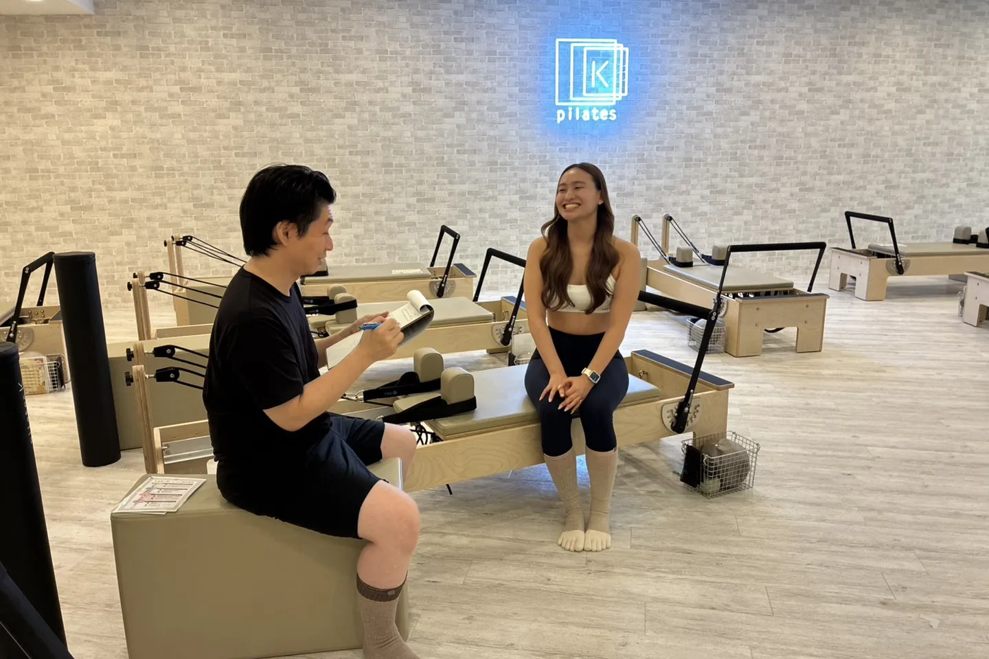 スタジオのリフォーマーに腰かけた maikaさんに、タカが話を聞いている。奥の壁に pilates K のロゴが青く光っている
