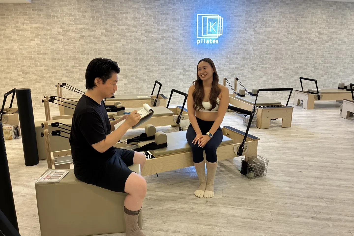 スタジオのリフォーマーに腰かけた maikaさんと、メモを手にしたタカが笑顔で話している。奥の壁に pilates K のロゴが青く光っている