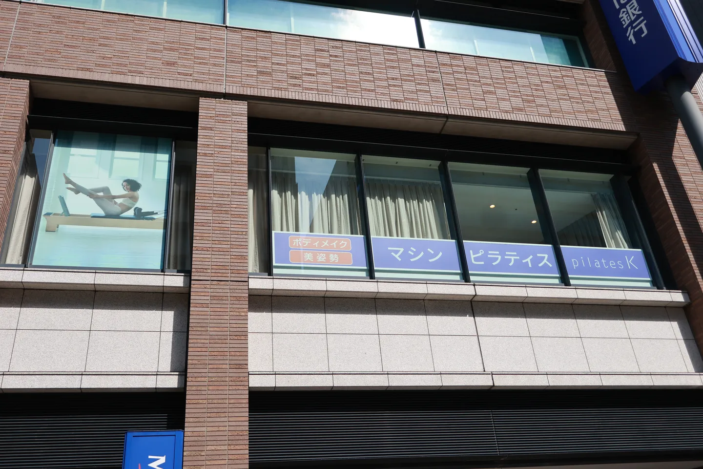 通りから見上げた2階の窓。「マシンピラティス」「pilates K」の表示と、リフォーマーで体を動かす写真が貼られている
