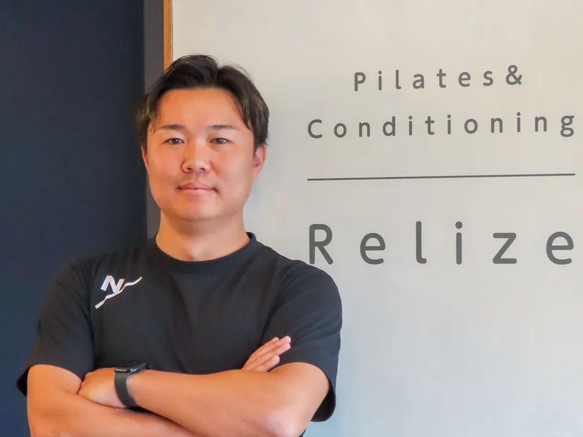 腕を組んで立つ吉成侑弥さん。後ろの壁に Pilates & Conditioning Relize のロゴがある